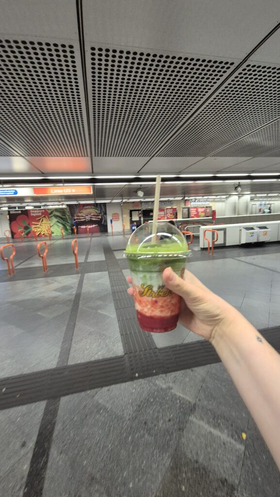 Hand hält einen To-go-Becher mit grünem und rotem Smoothie in einer U-Bahn-Station