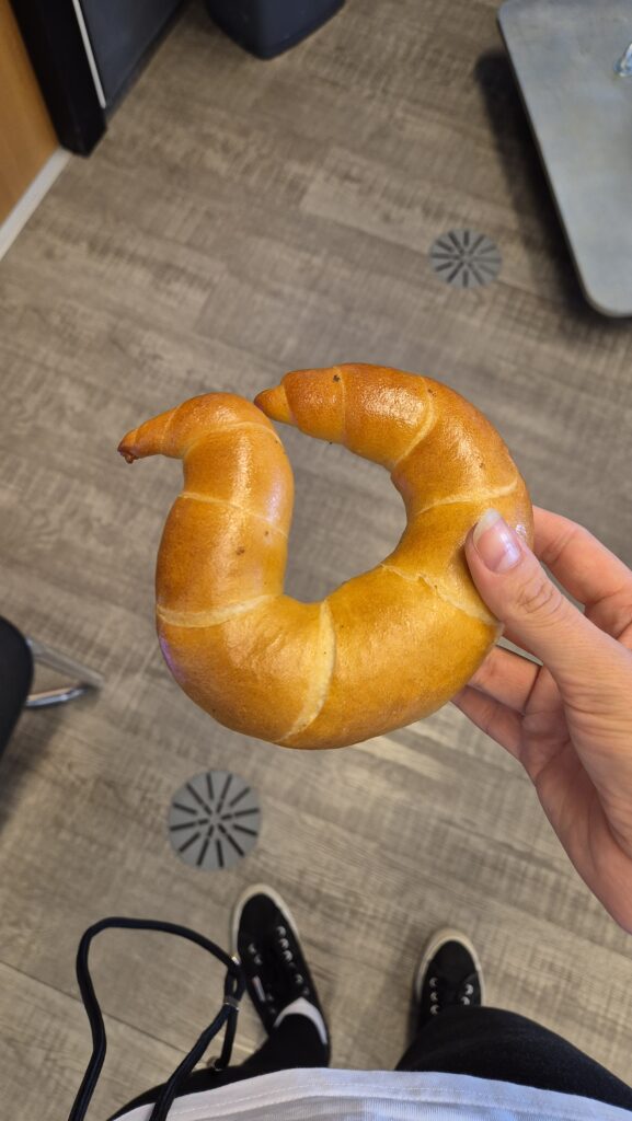 Hand hält ein großes goldgelbes Kipferl, im Hintergrund Boden und Schuhe